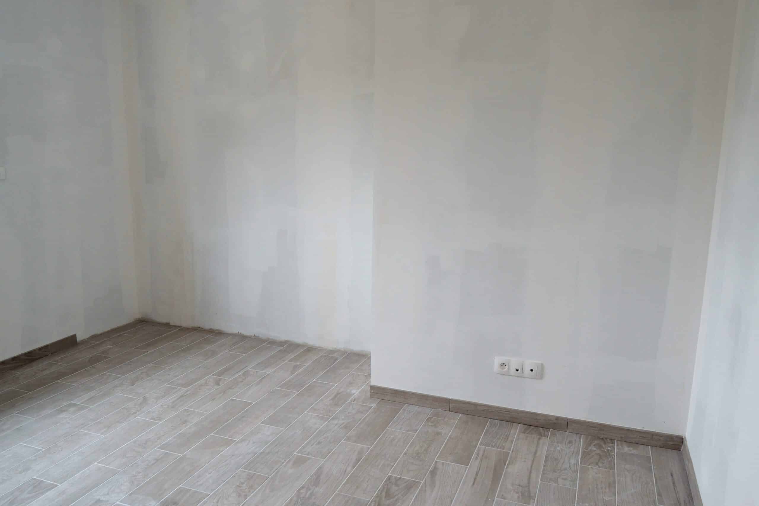 Pièce vide en rénovation avec murs blancs enduits et carrelage gris imitation bois posé au sol.